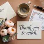 Flat lay of thank-you cards, pen, flowers, and coffee cup on wooden table for a thank-you guide